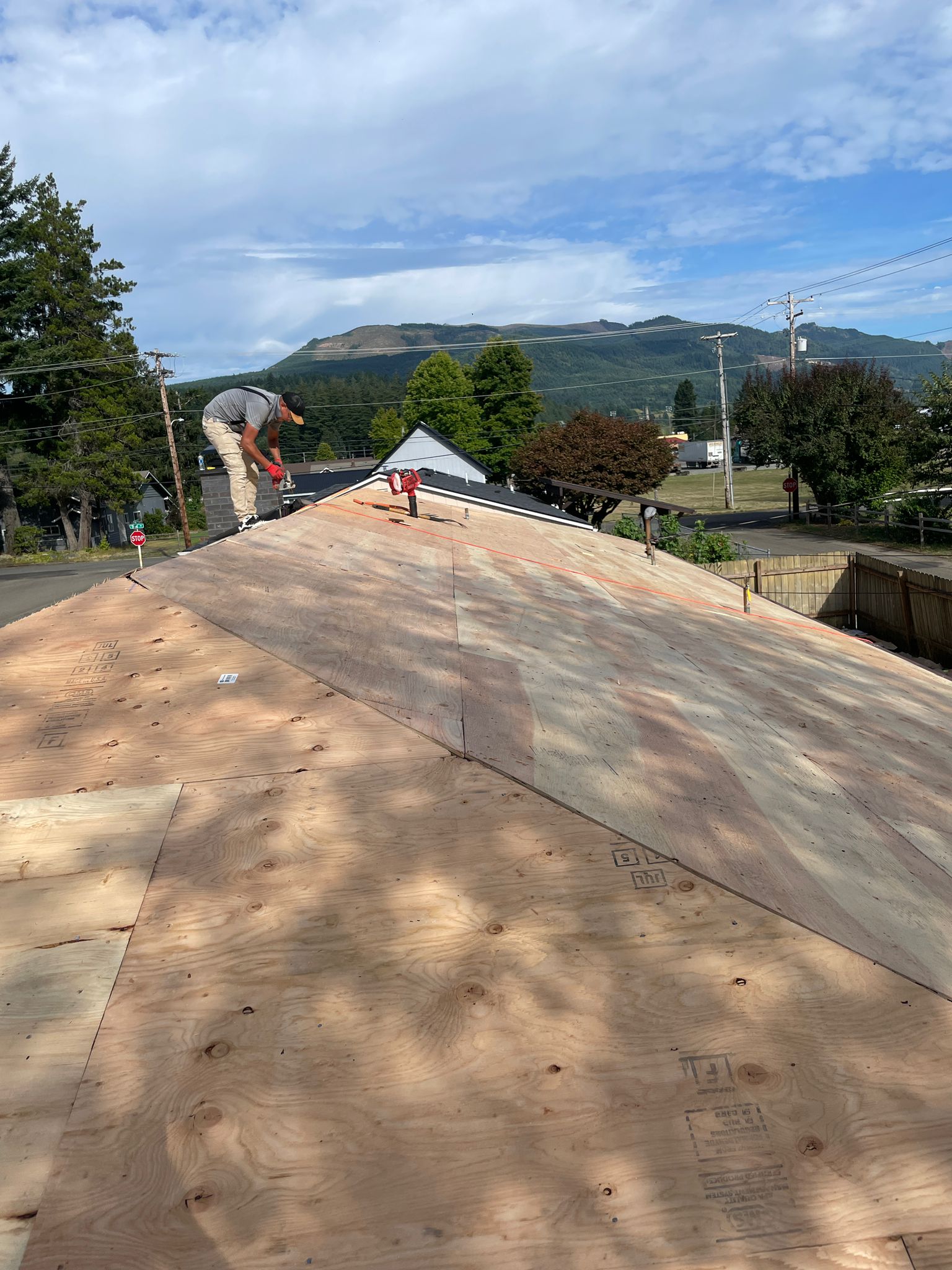 Roofer installing new plywood decking on a steep gable roof with mountain backdrop in Morton, WA, prior to Interlock Copper Penny aluminum slate application.