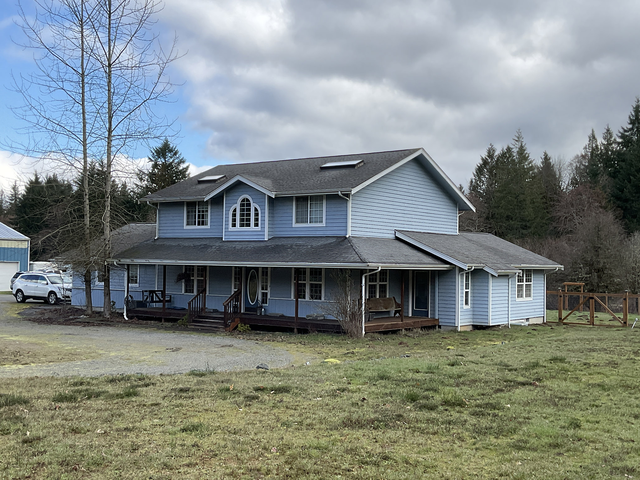 Two-story farmhouse-style home with deep charcoal Interlock cedar shingle metal roof, wrap-around porch, dormer, and skylights set on a rural forested acreage.