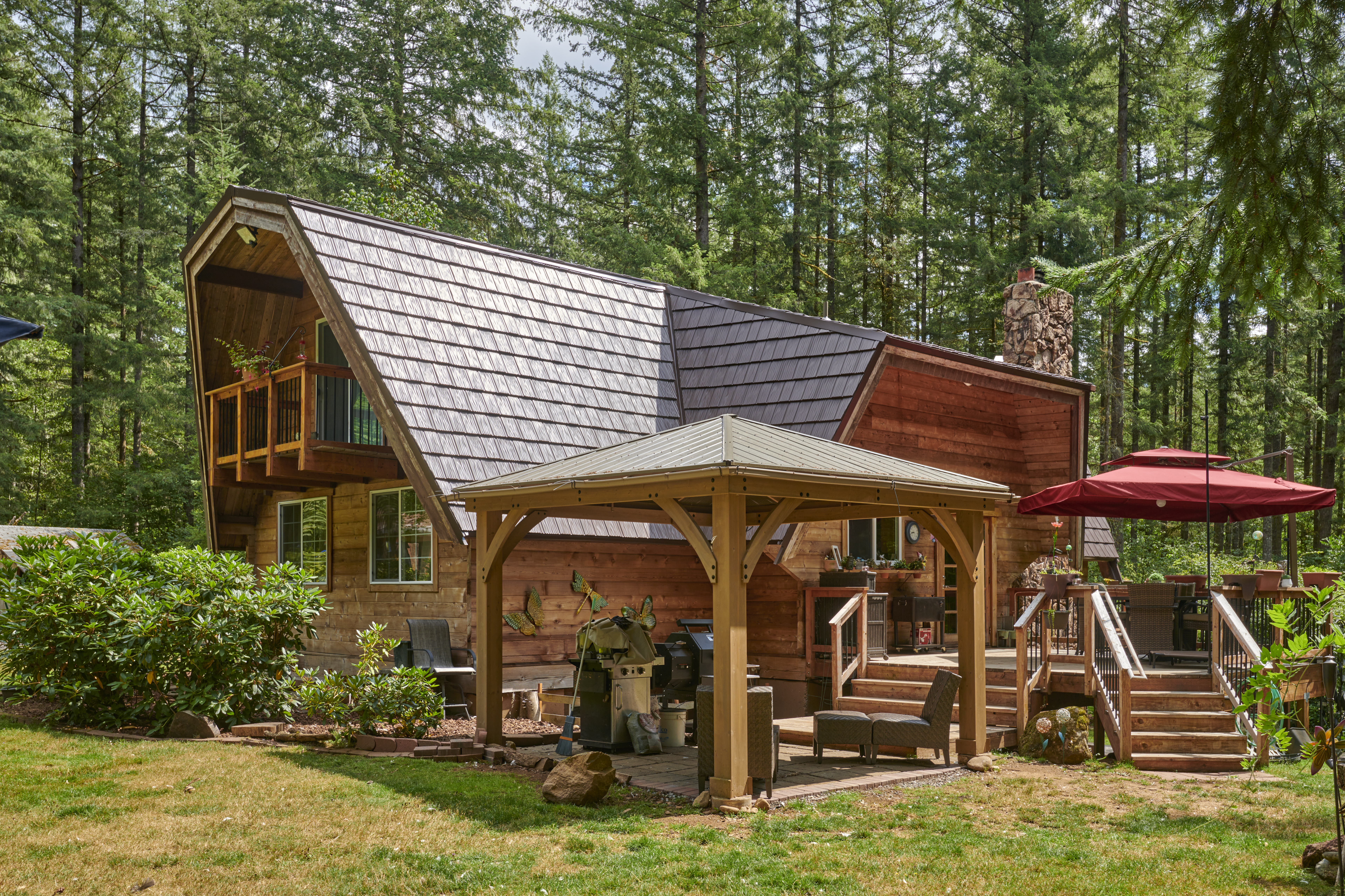 Rear-left view of a forested chalet-style home with steep Interlock aluminum shake roofing in Colonial Brown, stone chimney, upper balcony, and cedar siding.