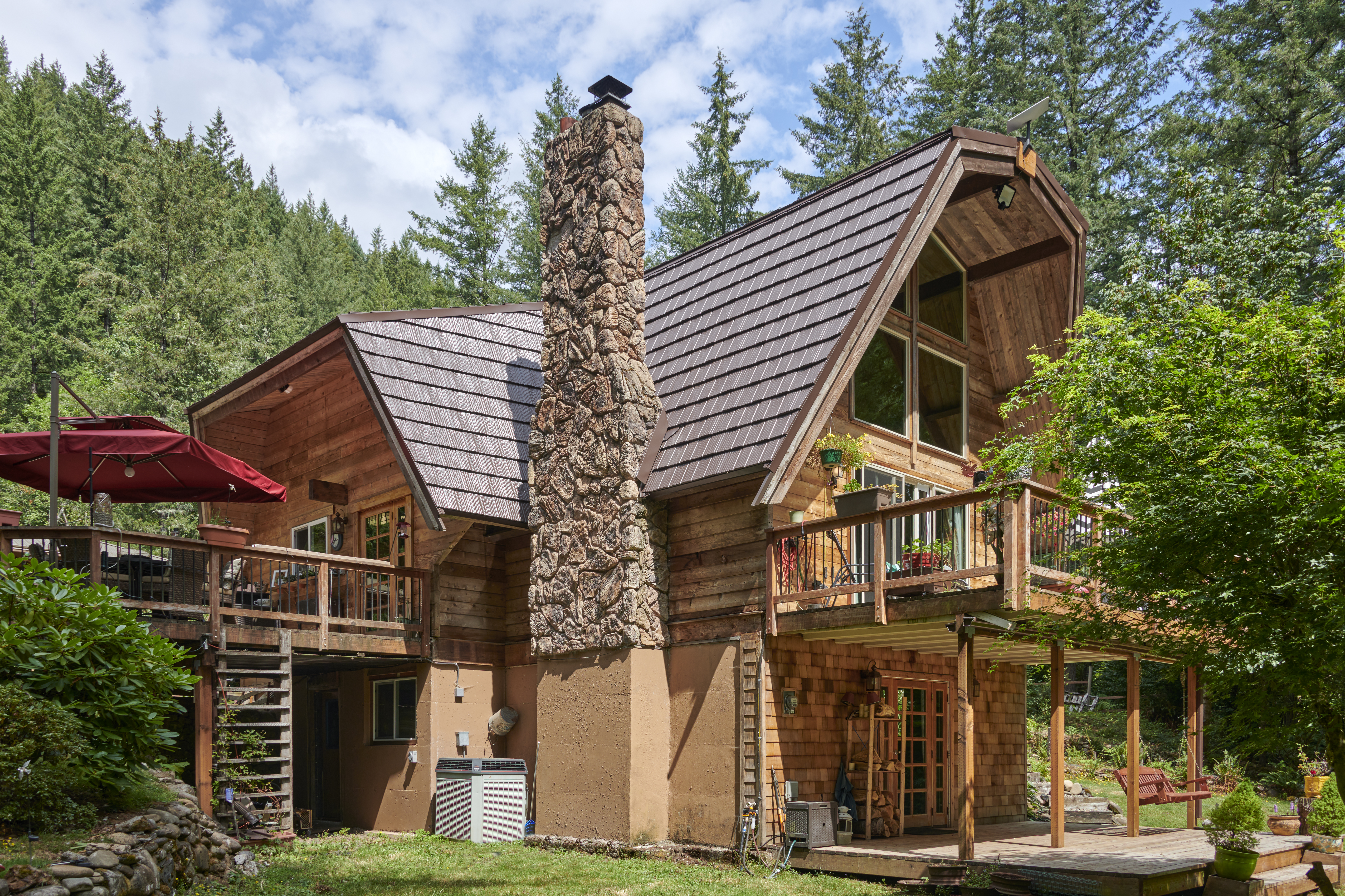 Rear-left view of a forested chalet with steep Colonial Brown Interlock aluminum shake roof, massive stone chimney, and multi-level wood decks.