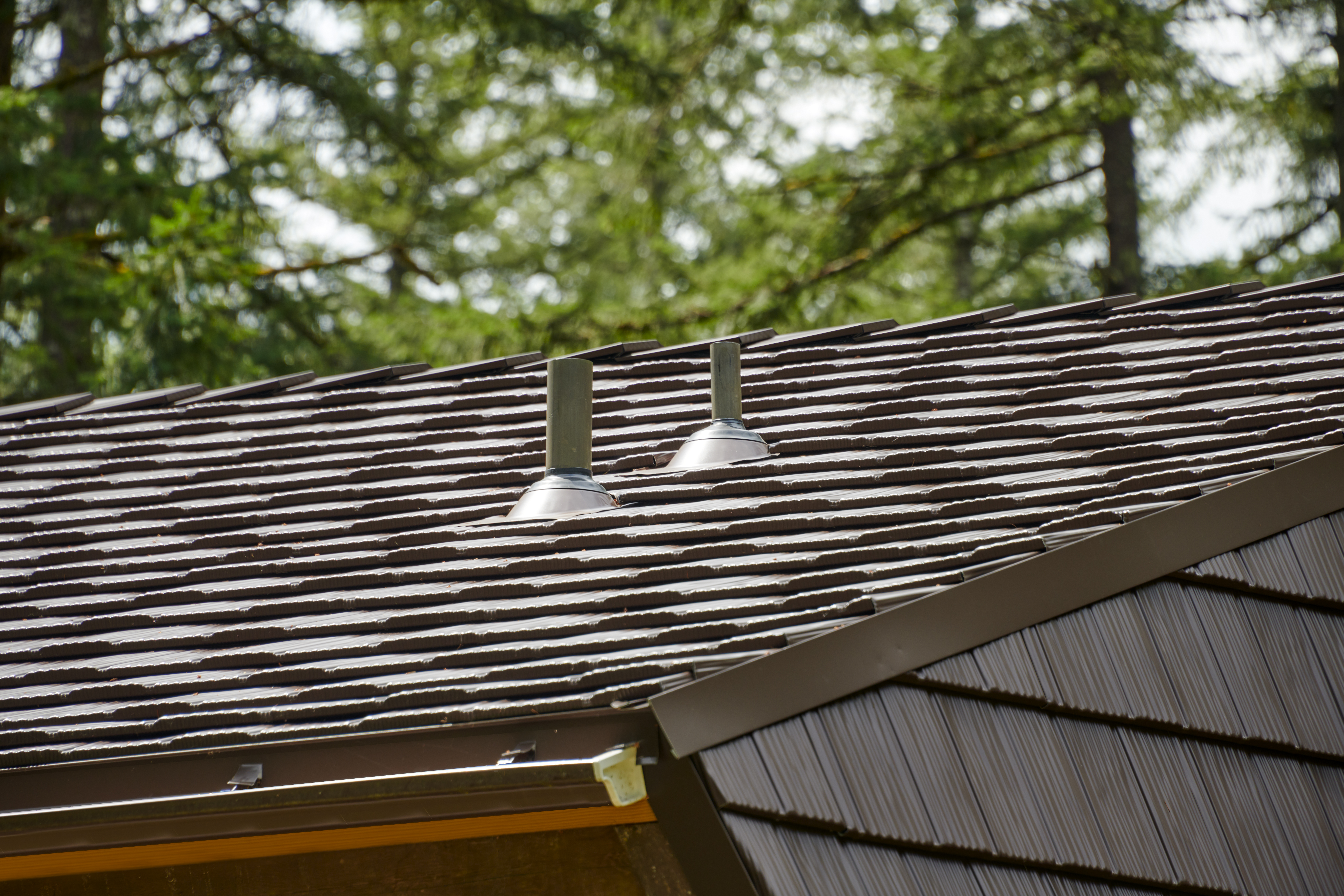 Close-up of two metal vent pipe flashings on a Colonial Brown Interlock aluminum shake roof with lush forested backdrop in Yacolt, WA.
