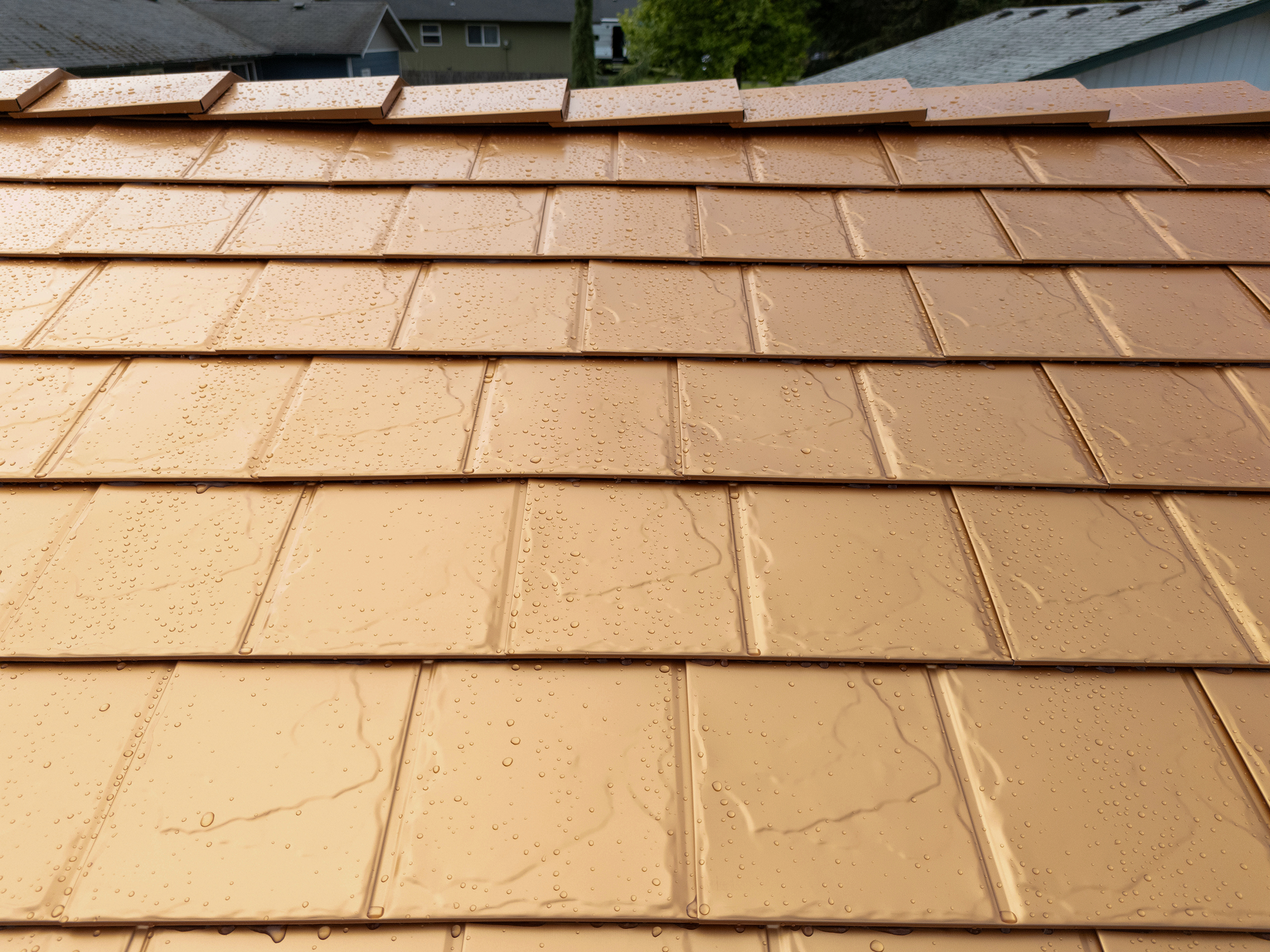 Close-up of Interlock aluminum slate metal roofing in Copper Penny finish, showing rain-beaded tiles with ridge cap detail against a suburban background.